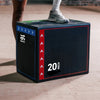 Person stepping onto a black and red exercise box with measurement labels on a wooden floor.