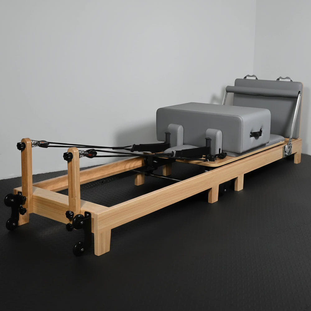 Pilates reformer with wooden frame and gray cushion on a dark floor.