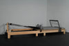 Wooden reformer exercise machine on a dark floor and gray wall background.