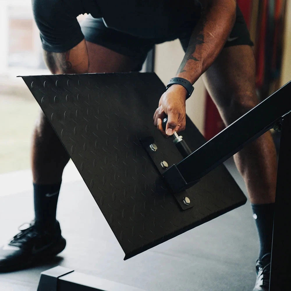 Person adjusting the foot plate of a black exercise machine in a gym setting