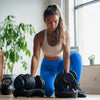 Woman in blue leggings and white sports bra lifting dumbbells in a home setting with plants and a window in the background.