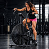 A woman riding an air bike exercise machine on a street, wearing a black tank top, pink shorts, and black athletic shoes.