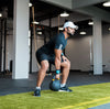 A man in a black athletic outfit and white cap exercises with a kettlebell on a green turf mat inside a gym.