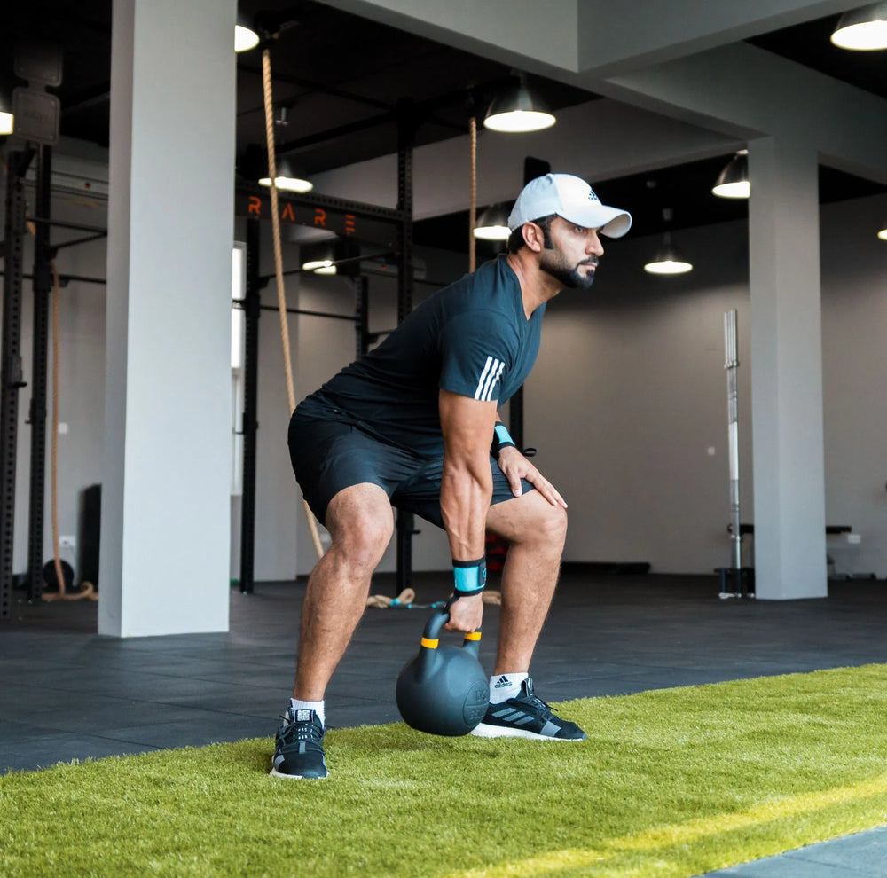 A man in a black athletic outfit and white cap exercises with a kettlebell on a green turf mat inside a gym.