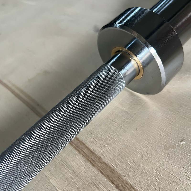 Close-up of a metal barbell with a knurled textured grip and polished steel ends, resting on a light wood surface.