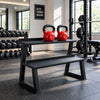 A black metal dual-tier bench with two red kettlebells on top, set on a black textured mat in a bright room with white cabinets and a window.