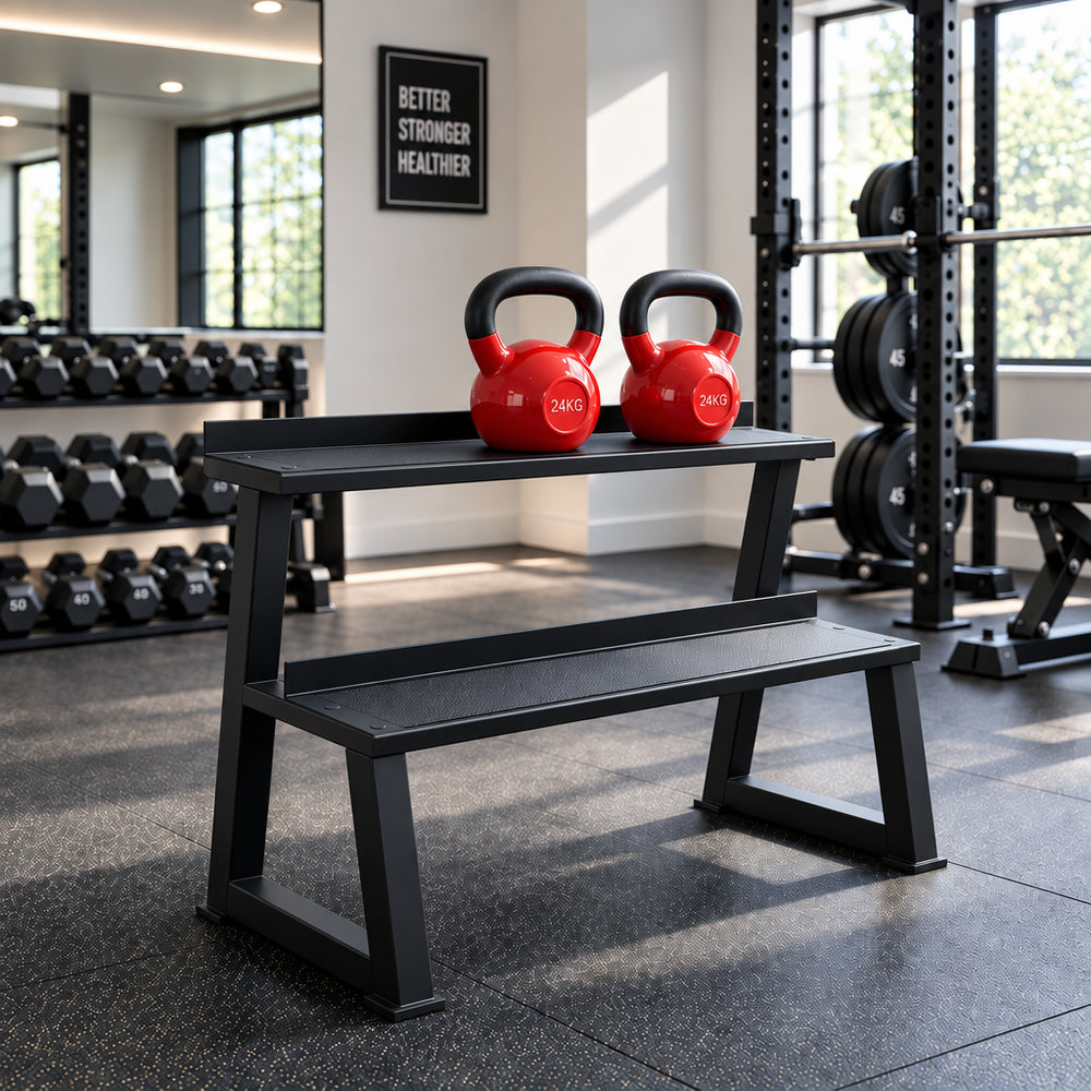 A black metal dual-tier bench with two red kettlebells on top, set on a black textured mat in a bright room with white cabinets and a window.
