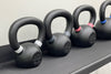Four black kettlebells on a dark textured surface against a light beige wall, with white, gray, pink, and blue bands around the handles and bases.