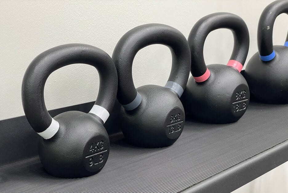Four black kettlebells on a dark textured surface against a light beige wall, with white, gray, pink, and blue bands around the handles and bases.