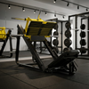 A black and orange leg extension gym machine with a barbell rack, shown on a clean light gray studio background.
