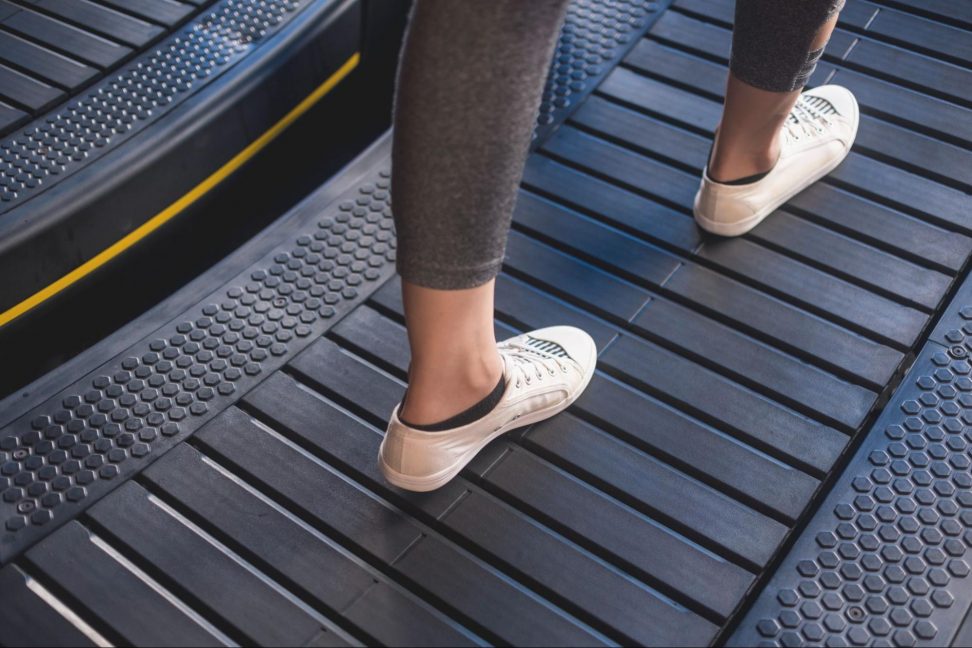 A close-up of a person's legs and feet wearing white slip-on sneakers with black trim, stepping onto a dark textured treadmill belt.