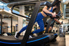 A woman in a blue sports bra and leggings runs on a treadmill inside a gym.