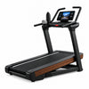 A black and wood-textured GTS Ultra treadmill with a digital display screen, positioned in a modern home gym setting with wooden floors, a bookshelf, and a large window showing green trees outside.