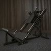 A gray adjustable leg press machine with a safety rail and weight stack, set on a light wood floor against a white wall with a brown curtain.