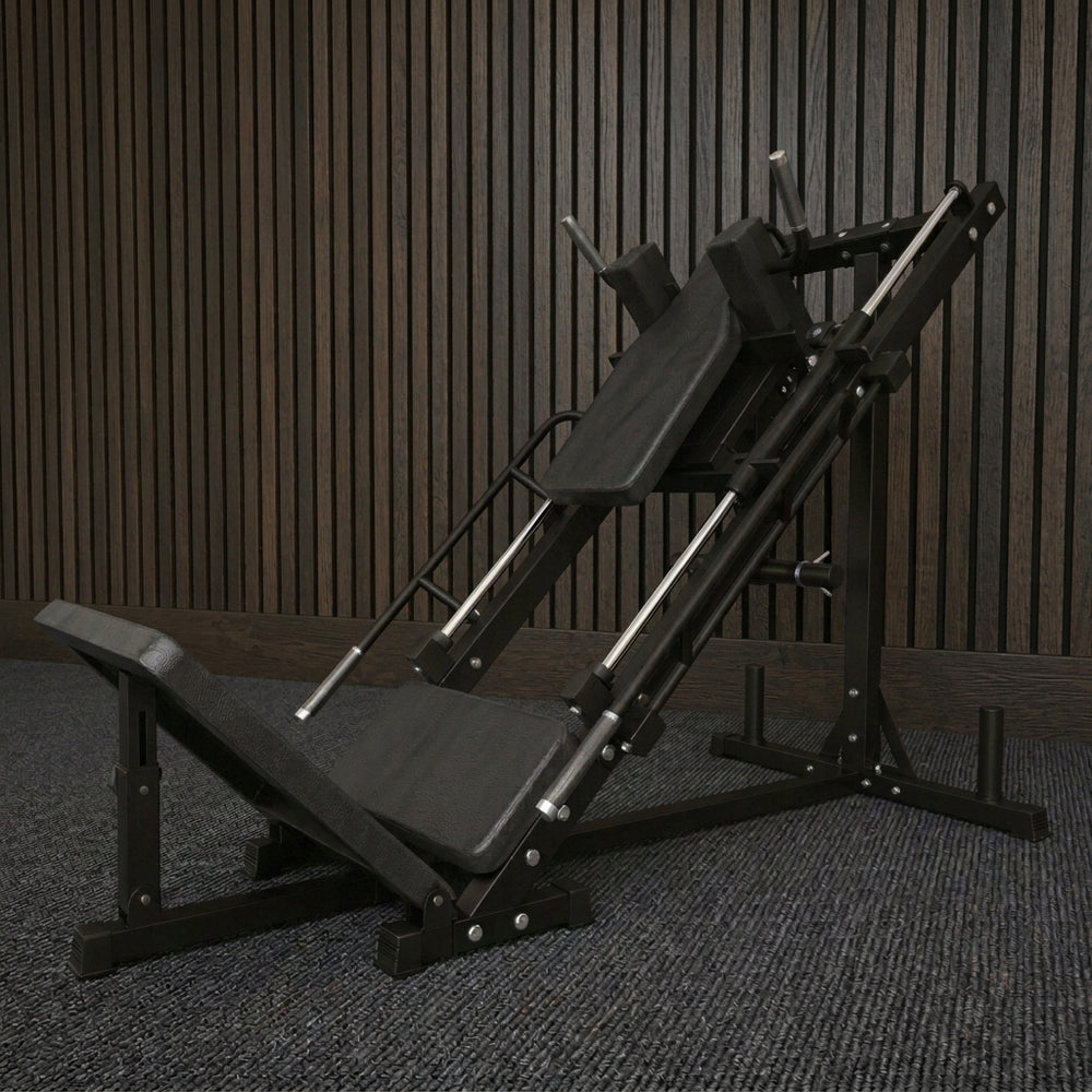 A gray adjustable leg press machine with a safety rail and weight stack, set on a light wood floor against a white wall with a brown curtain.