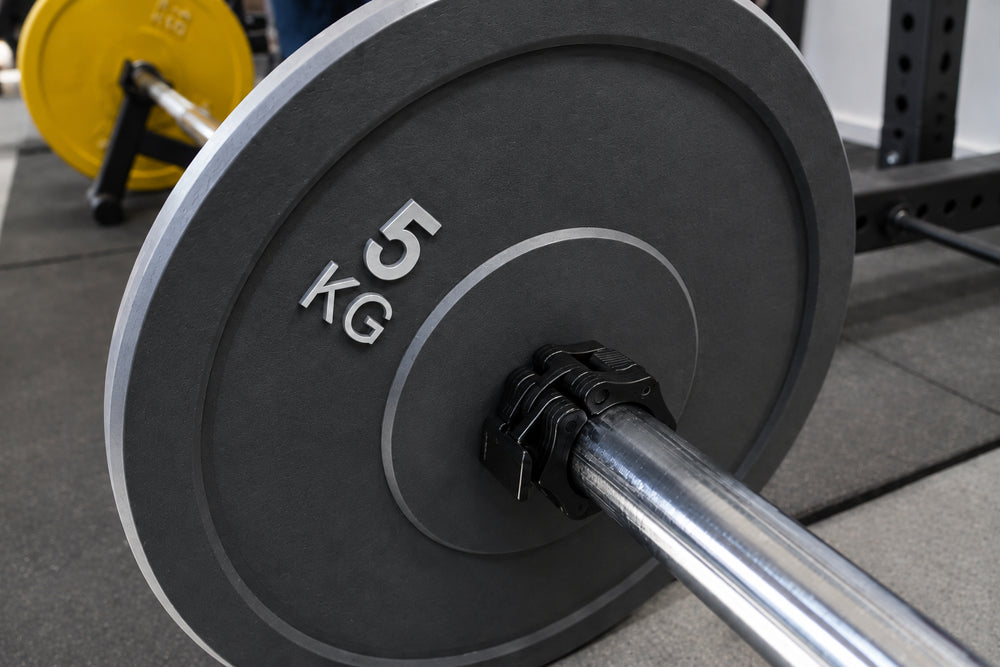 A close-up of a barbell with a large dark gray 5 kg weight plate, photographed in a gym setting.