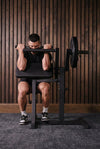 A man performing a shoulder press on a black gym bench press machine with a barbell loaded with weights, set against a dark gray wall.