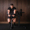 A man in a black t-shirt, black shorts, and a dark cap is performing a leg extension exercise on a black gym weight bench with a barbell weight plate.
